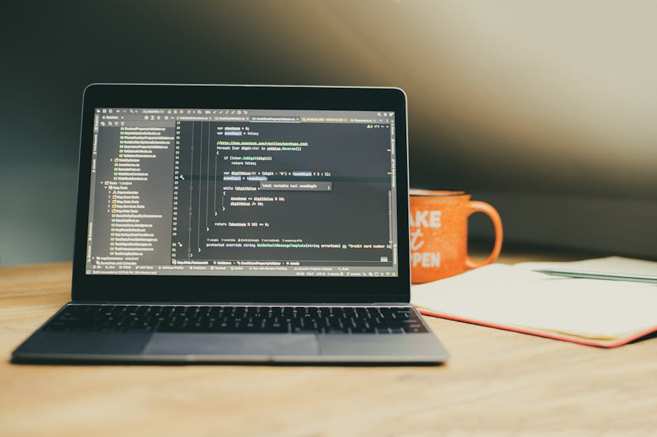 A close-up of a laptop displaying lines of computer code on a dark background with syntax highlighting. The laptop is placed on a wooden desk, accompanied by a partially visible orange coffee mug with