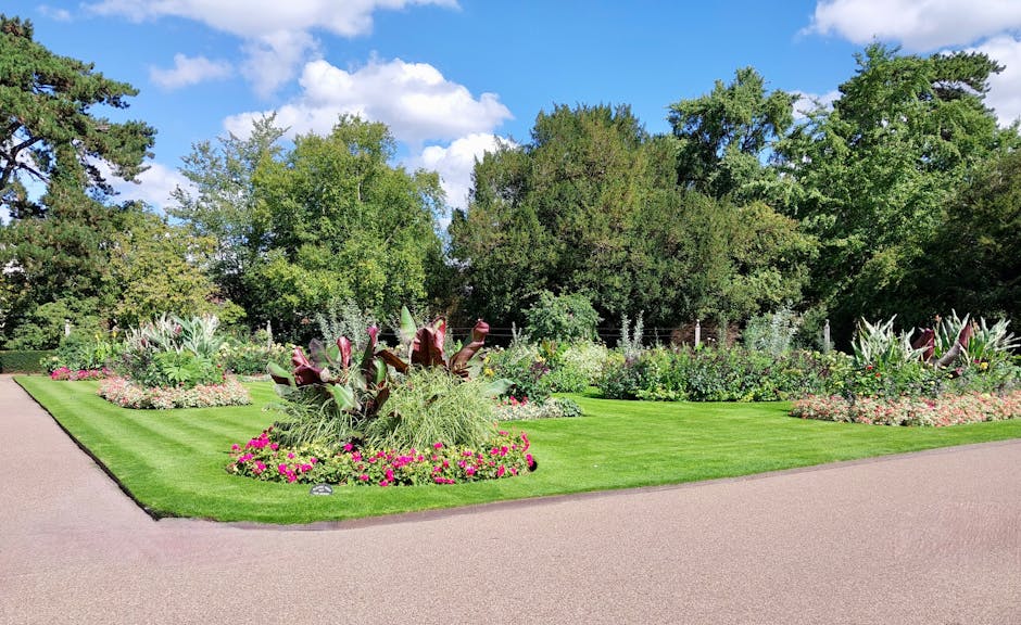 A well-maintained garden featuring a lush green lawn with neatly trimmed grass, bordered by vibrant flower beds filled with pink, white, and purple flowers. In the foreground, there is a circular flow