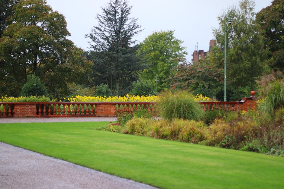 A well-maintained garden featuring a lush green lawn with neatly trimmed grass, bordered by vibrant flower beds filled with pink, white, and purple flowers. In the foreground, there is a circular flow