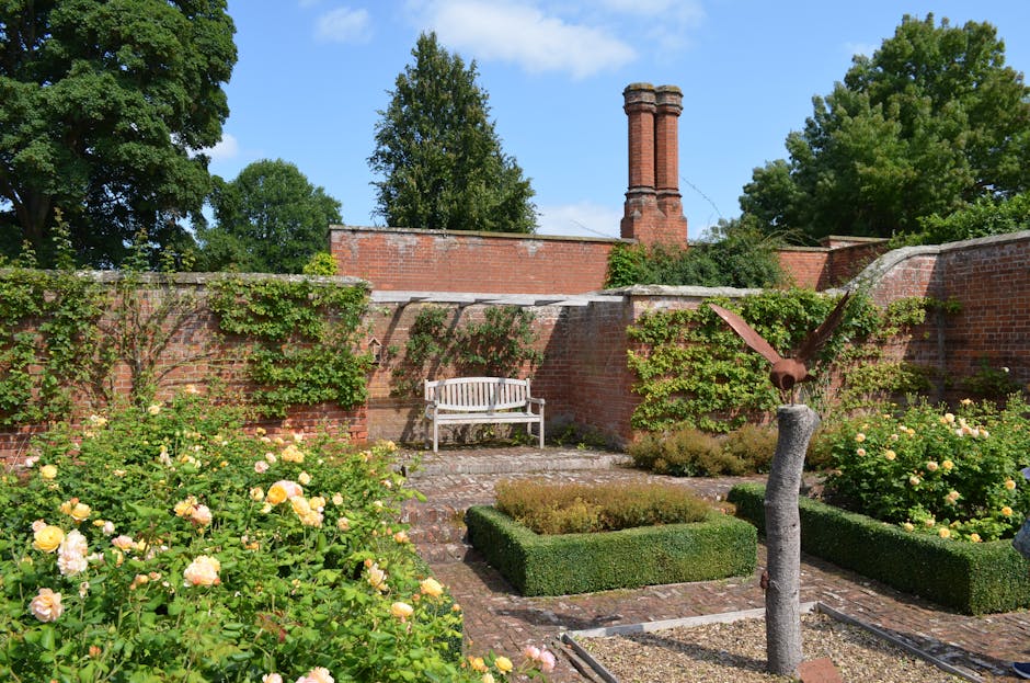 A historic red-brick building with numerous rectangular and circular windows and decorative architectural details in the background, behind a formal garden featuring a neatly maintained lawn, evenly s